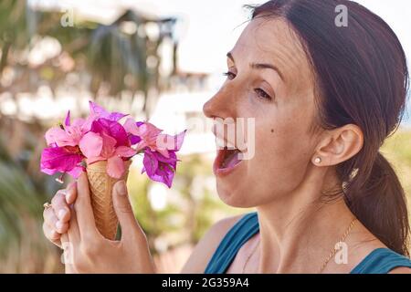 Jeune fille aux cheveux bruns essayant de manger de la glace avec des fleurs devant les feuilles de palmier par temps ensoleillé. Concept publicitaire de vacances d'été. Été surréaliste fleurs concept créatif et tendance Banque D'Images