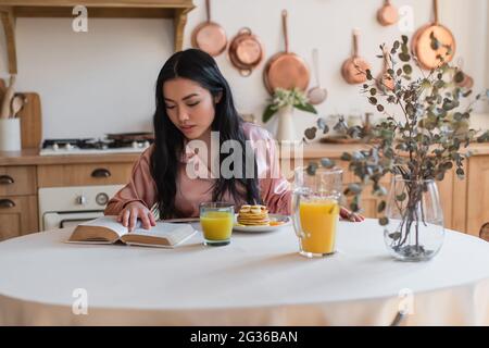 jeune fille asiatique en pyjama de soie lisant le livre tout en prenant le petit déjeuner dans la cuisine Banque D'Images