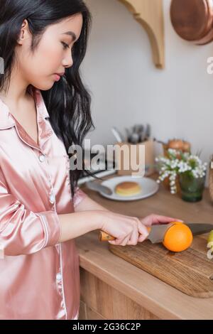 jeune femme asiatique en pyjama de soie rose coupant orange dans la cuisine Banque D'Images