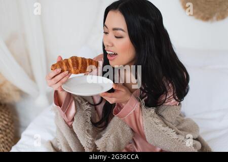 jeune femme asiatique en pyjama de soie et couverture assise sur le lit et en train de manger un croissant dans la chambre Banque D'Images