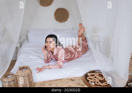 jeune femme asiatique en pyjama de soie rose couché sur le lit et parlant sur le téléphone portable dans la chambre Banque D'Images