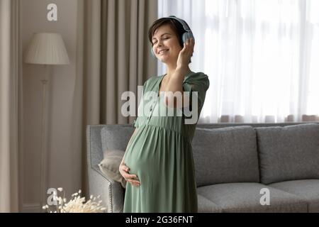 Femme enceinte souriante dans un casque qui profite de la musique préférée, touchant le ventre Banque D'Images