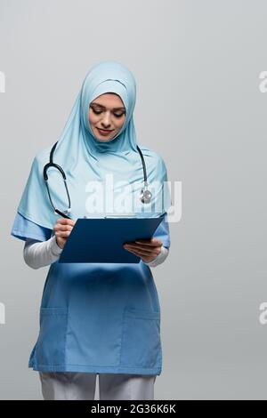 joli médecin musulman en hijab et écriture uniforme sur presse-papiers isolé sur gris Banque D'Images