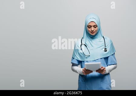 médecin arabe en hijab bleu et uniforme regardant la tablette numérique isolée sur gris Banque D'Images