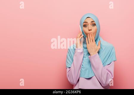 choqué femme arabe dans hijab bleu parlant sur téléphone portable sur fond rose Banque D'Images