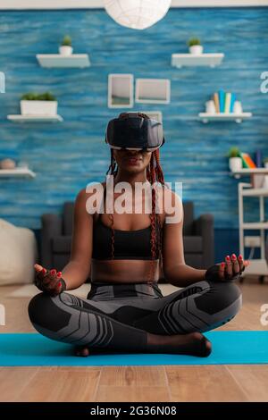 Femme noire expérimentant l'entraînement de réalité virtuelle corps et esprit méditant dans lotus pose assis sur le tapis de yoga dans la maison salon pour le calme, l'harmonie saine style de vie habillé dans des vêtements de sport. Banque D'Images