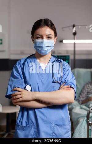 Portrait d'une infirmière médicale avec masque chirurgical dans la chambre d'hôpital avec malade unweel pantient senior avec traumatisme regardant la camea avec les bras croisés, portant des exfoliants. Banque D'Images