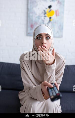 choqué femme musulmane couvrant la bouche avec la main tout en regardant la télévision à la maison Banque D'Images