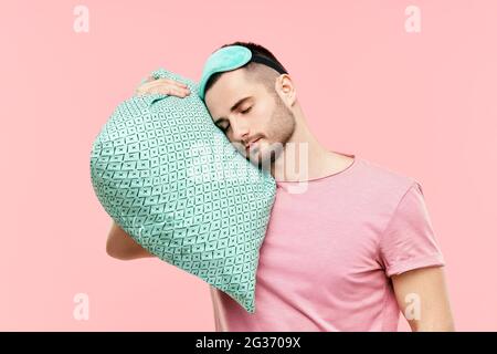 Jeune homme beau dormant tenant un oreiller dans les mains sur fond rose. Concept relaxant et fatigué Banque D'Images