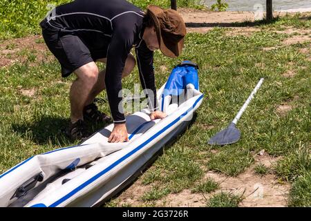 Homme d'âge moyen en chapeau marron, maillot de bain noir et short, pliant en kayak gonflable dégonflé. Préparation du stockage. Pagayez sur le sol à côté. Banque D'Images