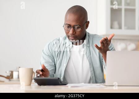 Concept d'épargne et de finances. Noir choqué mature homme calculant des factures, en utilisant la calculatrice, assis dans la cuisine Banque D'Images