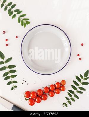 Composition verticale des aliments avec assiette blanche vide, tomates, herbes et épices sur une table blanche. Alimentation saine et concepts de nutrition alimentaire. Pose à plat Banque D'Images
