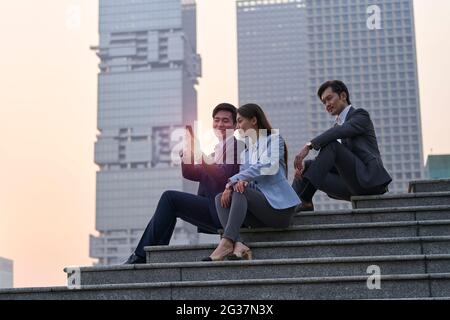 équipe de trois cadres d'entreprise asiatiques assis sur des marches regardant le téléphone mobile avec un fond de gratte-ciel Banque D'Images