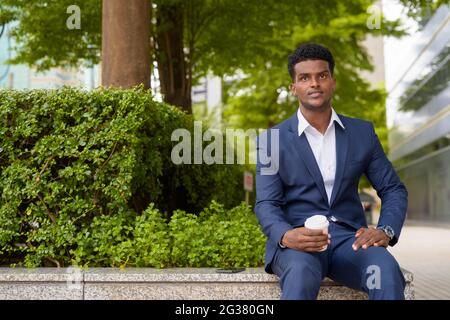 Portrait d'un jeune homme d'affaires africain tenant une tasse à café à emporter en plein air en ville tout en étant assis à l'horizontale Banque D'Images