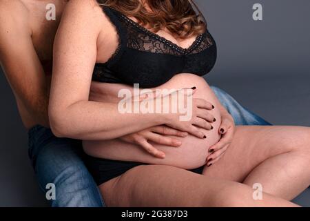 S'asseoir femme enceinte tient les mains avec son mari dans le grand ventre. Couple s'attendant à un bébé dans un fond gris. Banque D'Images