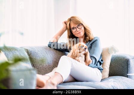 Photo d'une jolie femme d'âge moyen qui se détend sur le canapé tandis que son adorable chiot cavalier est assis sur ses genoux. Banque D'Images