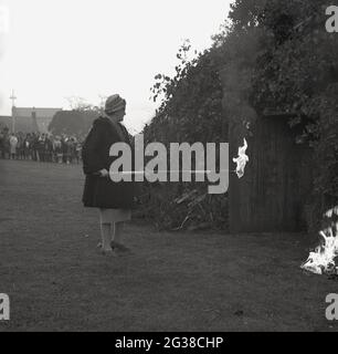 1960s, historique, soirée dans un parc et une dame âgée dans un manteau et un chapeau de fourrure courts, Et tenant un long poteau éclairé à une extrémité avec une flamme sur le point d'entrer dans l'entrée d'un grand feu de camp couvert de branches d'arbres pour mettre le feu il s'allume, Fife, Écosse, Royaume-Uni. Banque D'Images