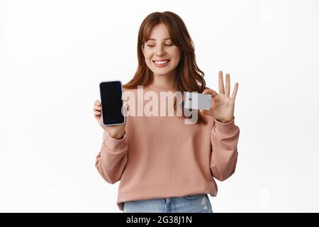 Souriante fille rouge montre carte de crédit et application de téléphone mobile, regarde l'écran de smartphone, se tient dans des vêtements décontractés sur fond blanc Banque D'Images