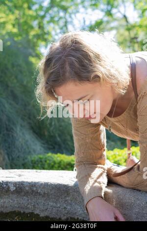 Belle femme blonde d'âge moyen penchée et penchée avec ses coudes sur le bord d'un puits d'eau et regardant à l'intérieur Banque D'Images