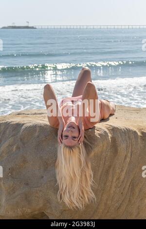 Belle mannequin à cheveux blonds couchée à l'envers sur un rocher surplombant les vagues avec les mains sur ses joues tandis que les cheveux pendaient. Banque D'Images