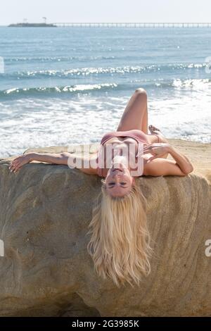 Magnifique modèle féminin à cheveux blonds couché à l'envers sur un rocher surplombant les vagues avec les yeux ouverts tandis que les cheveux pendent. Banque D'Images