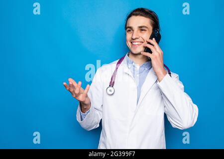 Photo de heureux médecin portant blouse blanche et stéthoscope talking on cell phone isolés sur fond bleu Banque D'Images