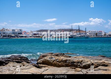 Mykonos, Cyclades, Grèce. 21 mai 2021. Maisons blanchies à la chaux bâtiments commerces sous ciel bleu à côté de la mer Egée arrière-plan bateaux au port. Été célèbre Banque D'Images