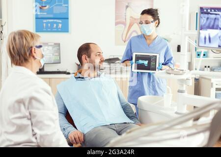 Assistant de dentiste montrant la radiographie des dents sur l'écran d'ordinateur de tablette à l'homme malade de la clinique dentaire. Orthodontiste discutant du traitement des maux de dents pour prévenir les problèmes de stomatologie Banque D'Images