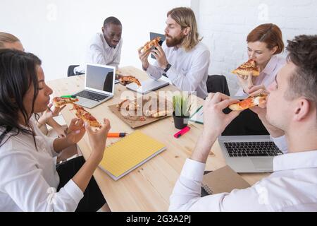 Un groupe de gens d'affaires divers qui mangent de la pizza en pause tout en travaillant ensemble dans un bureau assis autour du bureau Banque D'Images
