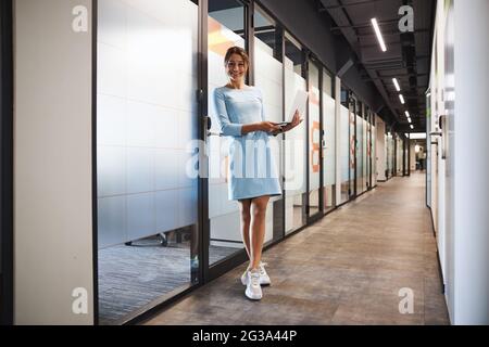 Belle jeune femme caucasienne entrepreneur en robe bleue tenant l'ordinateur portable dans les mains dans le bureau de démarrage Banque D'Images