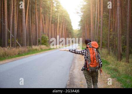 Une touriste féminine dans une chemise à carreaux avec un grand sac à dos orange près d'une autoroute dans les bois vote pour obtenir un tour. Randonnée pédestre, tourisme domestique. Backpa Banque D'Images