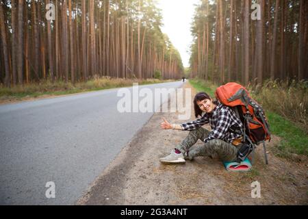 Une touriste féminine dans une chemise à carreaux avec un grand sac à dos orange près d'une autoroute dans les bois vote pour obtenir un tour. Randonnée pédestre, tourisme domestique. Backpa Banque D'Images