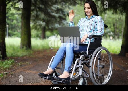 Une jeune femme handicapée en fauteuil roulant agite la main à l'écran d'ordinateur portable dans le parc Banque D'Images