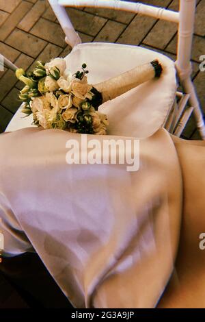 Vue de dessus de la jambe d'une femme dans une robe blanche soyeuse sur une chaise de mariage avec un bouquet de roses blanches Banque D'Images