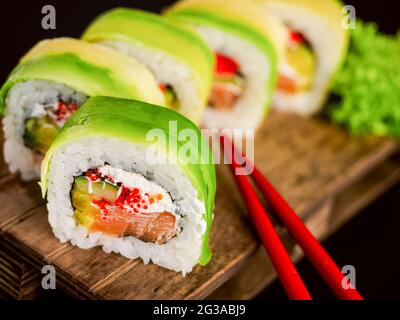 Les sushis roulent de l'avocat avec du saumon, de la crème de philadelphie, du tobiko et du concombre sur bois, des baguettes rouges et sur fond noir. Sushi moi Banque D'Images