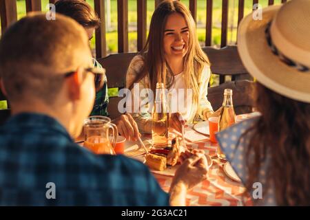 Les jeunes s'amusent sur la terrasse, boivent des boissons et bavardez pendant que leurs amis servent de la viande grillée sur un barbecue à l'extérieur au coucher du soleil Banque D'Images