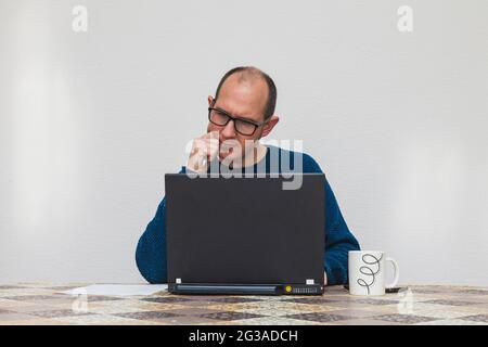 Un homme portant des lunettes et un jersey bleu est pensif devant un ordinateur portable. Sur la table il y a une tasse blanche, un smartphone, une feuille de papier et un stylo. Banque D'Images