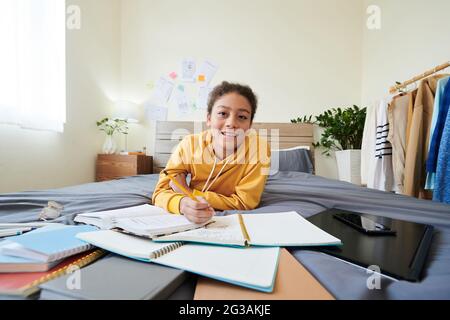 Portrait d'une jeune fille d'école secondaire de race mixte, couchée sur un lit parmi les cahiers de travail dans sa propre chambre et prenant des notes tout en faisant des devoirs Banque D'Images
