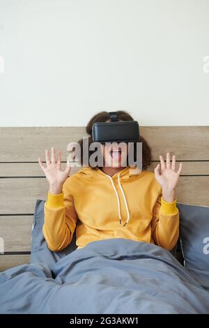 Adolescente de course mixte excitée dans un chandail jaune assis sous une couette et tenant les mains vers le haut tout en hurler à l'aide d'un simulateur de réalité virtuelle Banque D'Images
