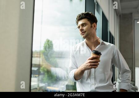 homme d'affaires positif et rêveur debout près de la fenêtre avec un café à emporter Banque D'Images
