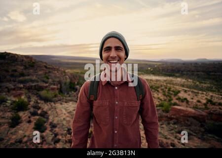 Portrait d'un randonneur homme heureux réussi avec sac à dos et casquette regardant l'appareil photo Banque D'Images