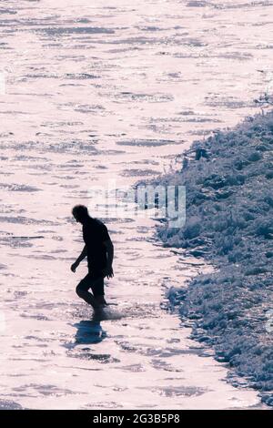 Un homme silhoueté par la lumière du soir qui sort de la mer, suivi d'une petite vague qui coule doucement sur la plage de Fistral à Newquay, dans les Cornouailles. Banque D'Images