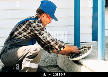 Une vieille couche de briques pose le porche d'un bâtiment en carreaux de béton sur la rue, un jour d'été. Un travailleur âgé en salopette effectue une tâche. Banque D'Images