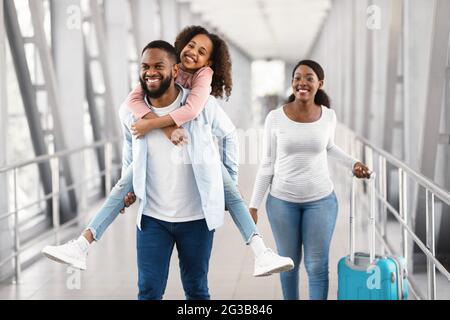 Bonne famille à l'aéroport. Portrait d'un homme noir à cheval sur sa fille à l'arrière, femme souriante marchant avec une valise dans le hall de l'aérogare. Papa qui donne des gi Banque D'Images