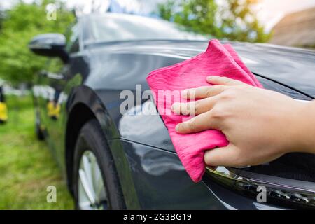 Une main avec un chiffon rouge essuie les gouttes d'eau sur une voiture après lavage avec du shampooing et de la cire de voiture dans le pays. Laver à la main Banque D'Images