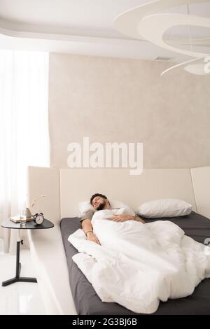 Un homme reposant dormant sous des draps blancs dans un grand lit Banque D'Images