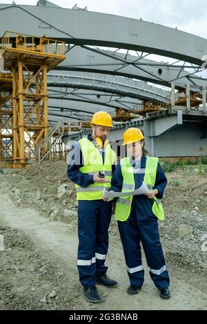 Deux constructeurs en uniforme regardant l'esquisse sur le plan tout en discutant Banque D'Images