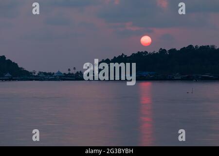 Un immense soleil, teinté de rouge, se trouve derrière la forêt tropicale qui entoure la petite ville de Bang Bao, sur l'île de Koh Chang, golfe de Thaïlande Banque D'Images