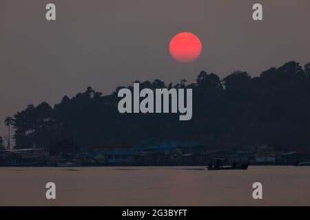 Un immense soleil, teinté de rouge, se trouve derrière la forêt tropicale qui entoure la petite ville de Bang Bao, sur l'île de Koh Chang, golfe de Thaïlande Banque D'Images