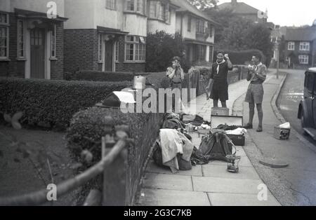 1951, historique, nous l'avons fait!... à l'extérieur dans une rue de banlieue, par une voiture de l'époque, deux maîtres scouts et un jeune scout, se tiennent près de leurs bagages et matériel de camping, pots et casseroles qui sont situés sur le trottoir. L'un des hommes lève une pompe manuelle en hommage à la fin réussie de leur voyage au camp de scouts, Angleterre, Royaume-Uni. Banque D'Images